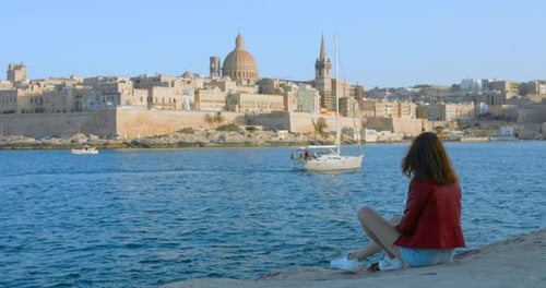A woman sitting and reading near the shore with the city of Valletta in the horizon.