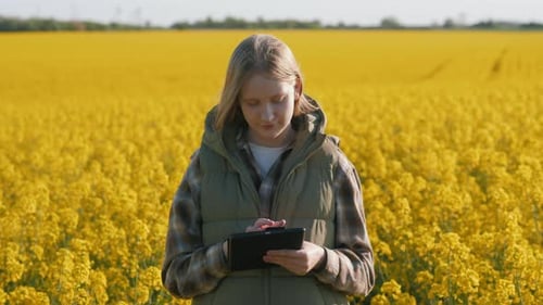 A Teenage Girl Uses a Tablet Against the Backdrop of a Field of Blooming Rapeseed