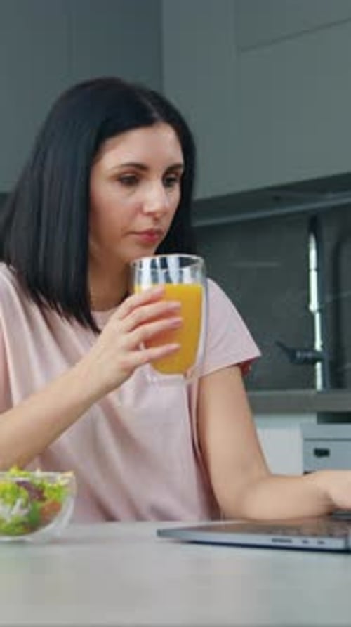 Woman Drinking Juice and Using Laptop in Kitchen