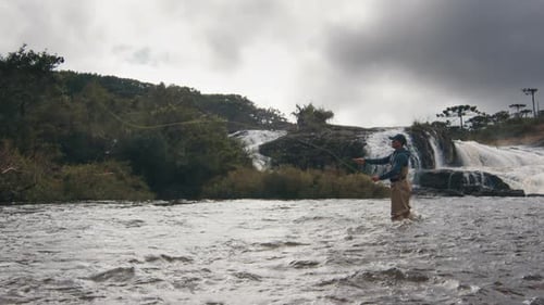 Fisherman Wading in River With Lush Waterfall