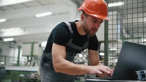 Young worker in uniform stands in the factory and typing on the laptop