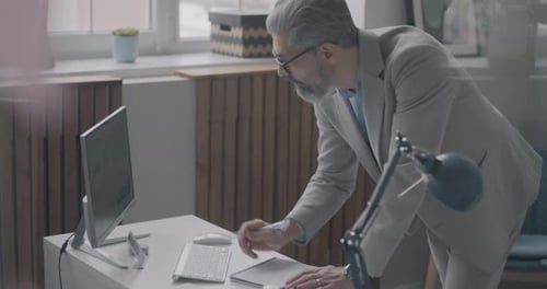 Mature Businessman Writing in Notebook Looking at Computer Screen Working in Office