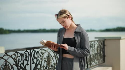 Focused Young Lady in Grey Reading Book As Wind Blows Pages By River
