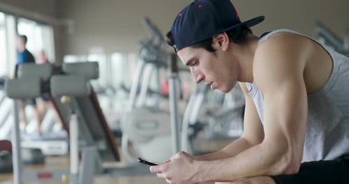Portrait of a guy in a gym while resting between a workout and the other uses his phone to watch s