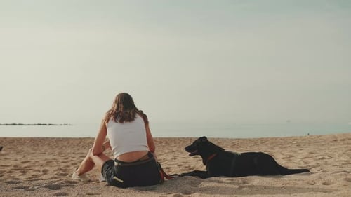 Girl with long dark blond wavy hair sits on the sand on the beach with black dog