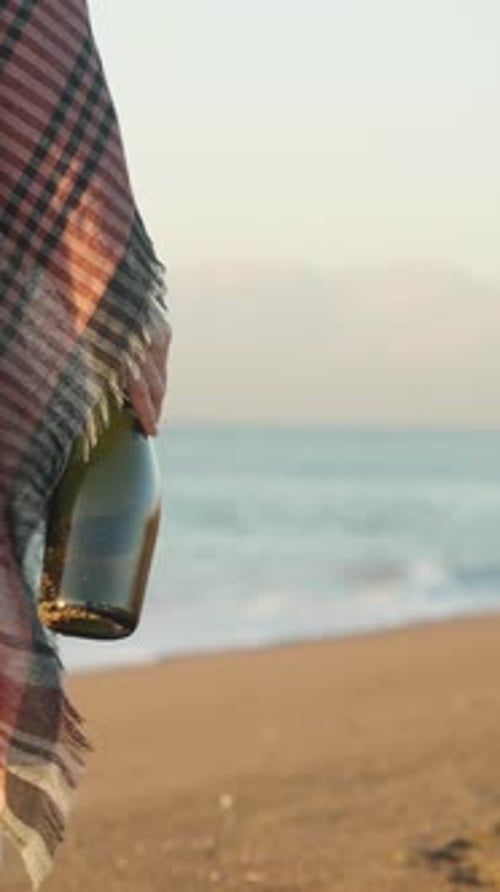 Vertical. At sunrise, a young woman walks along the beach with a bottle of champagne in her hand.