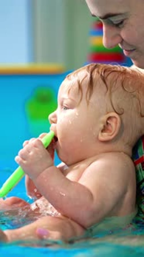 Woman is holding a little cute baby boy in the swimming pool. Lovely kid is chewing the toy.