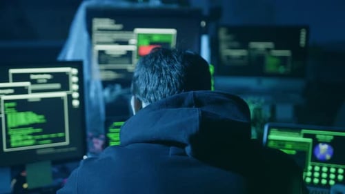 Un hacker sigiloso en una habitación con poca luz rodeado de múltiples pantallas y equipos de computadora