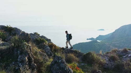 Young Man Hiking Along Rocky Mountain Top with Backpack Against Sky