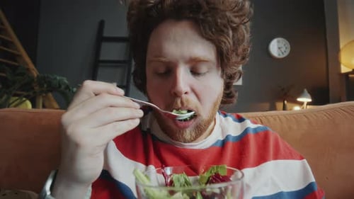Young Man with Obesity Eating Healthy Green Salad on Couch at Home