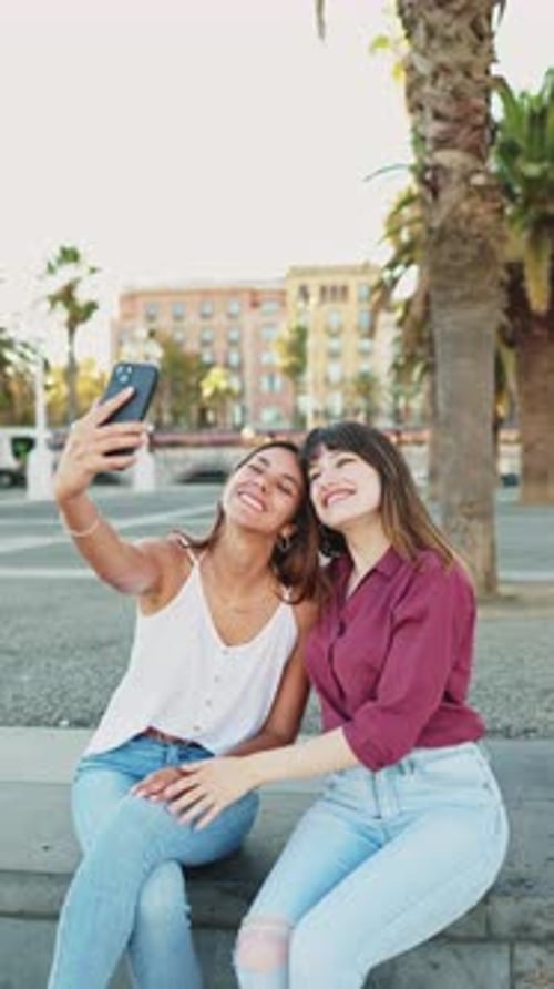 Two friends sitting on a bench laughing take a selfie with their smartphones to post on social media