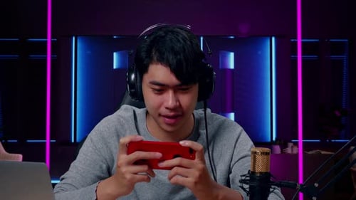 Close Up Of Smiling Asian Man Gamer With Computer Using Mobile Phone Playing Game In Neon Room