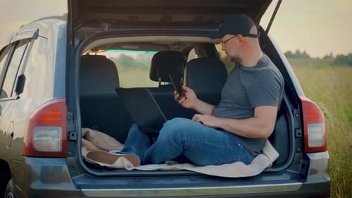 Homem olhando para a tela do smartphone trabalhando no teclado do laptop sentado no porta-malas do carro enquanto viaja