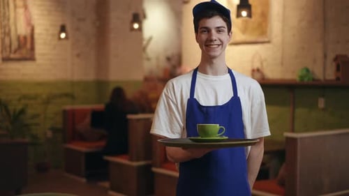 A Guy Waiter in a Blue Apron and a Blue Baseball Cap Holds a Tray with a Green Saucer and a Cup and