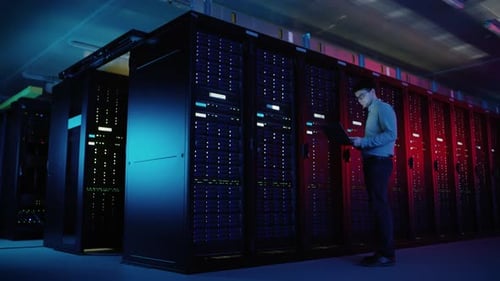 In Data Center: Male IT Engineer Walks along the Row of Operational Server Racks, Uses Laptop for M
