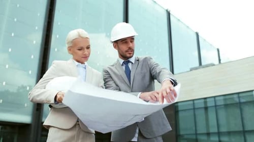 Smiling Businessman and Businesswoman with Blueprint and Helmets Pointing Finger on City