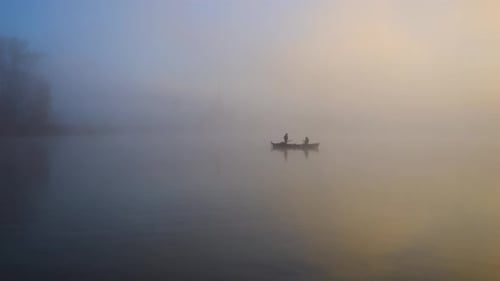 Two fishermen, whose silhouette can be seen, are on the lake in fog and sunrise