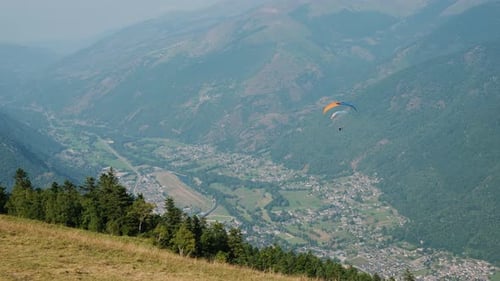 Paragliders soar over a lush valley with a distant plane below, surrounded by scenic mountains