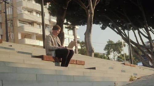 A Young Man is Working on a Laptop on a Bench on the Steps During Sunrise