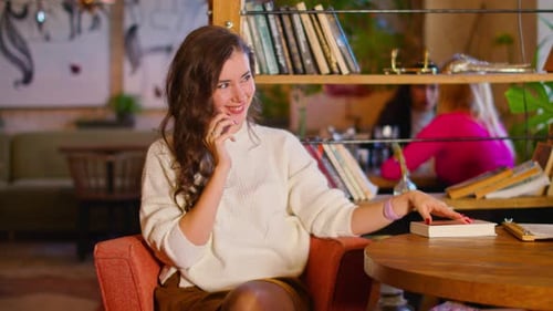 Happy Young Woman Speaks on the Phone in a Cafe Touches a Book with Her Hand