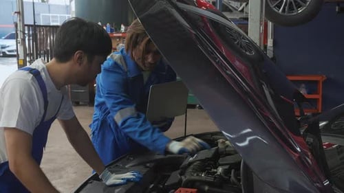 Two young man is mechanic using laptop computer checking engine of car in the garage.