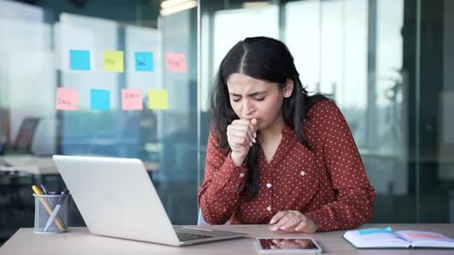 Sick young businesswoman coughing while sitting at a workplace in a modern business office. Tired