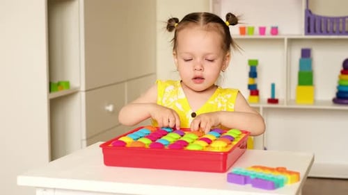 A two-year-old girl collects colorful mosaic tiles from the playing field.
