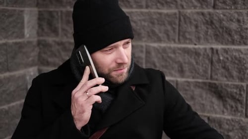 Handsome Man Wearing Black Coat and Brown Leather Bag Walking Down on the City with Mobile Phone in