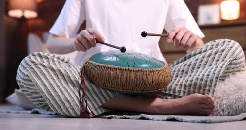 Woman playing steel tongue drum with mallets on floor at home, closeup