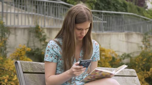 A Beautiful Young Caucasian Woman Works on a Smartphone and Reads a Book As She Sits on a Bench