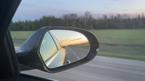 Reflexion im Autospiegel während der Fahrt bei Sonnenuntergang auf einer langen leeren Straße in ländlicher Gegend - am späten Abend