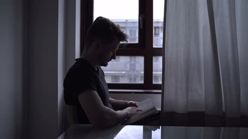 Young Man Reading Book by Window in Dim Light