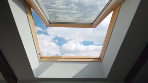 Looking on clouds flying past at the blue sky through open attic window in house roof.