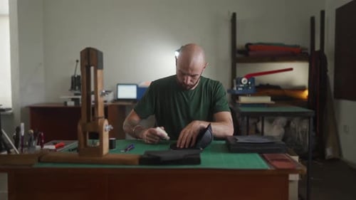 Un jeune artisan en cuir professionnel est assis à la table de travail et fabrique un sac à main noir dans un atelier Bold Man