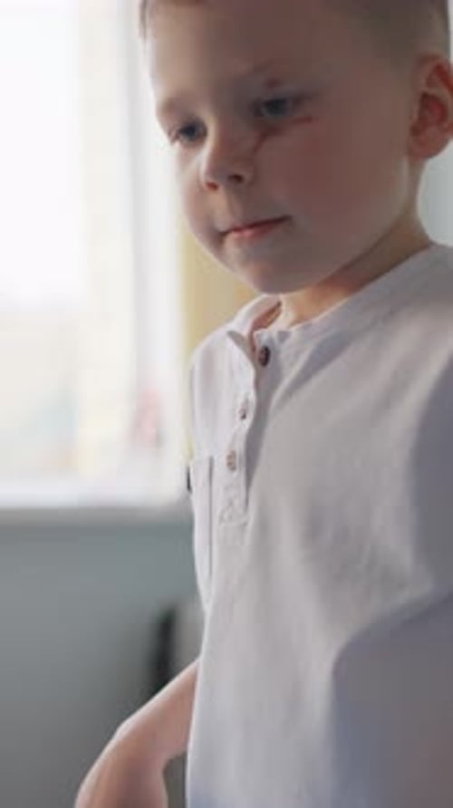 Young Boy in a White Shirt Standing Indoors Near a Window Interacting with a Red Cup on a Table