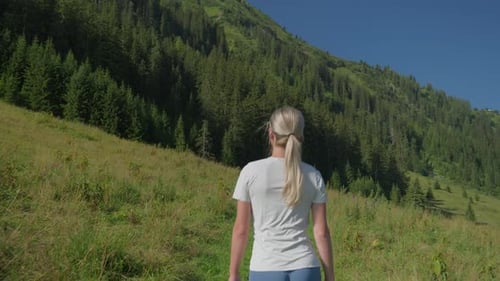 A woman hikes through a green mountain meadow under a clear blue sky on a sunny day