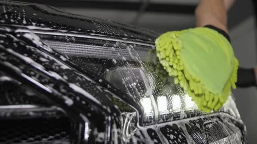 A Worker's Hand with a Green Sponge in Foam Washes the Car From the Front