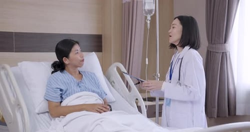 Slowmotion scene showing asian female doctor talking to female patient lying in hospital bed during