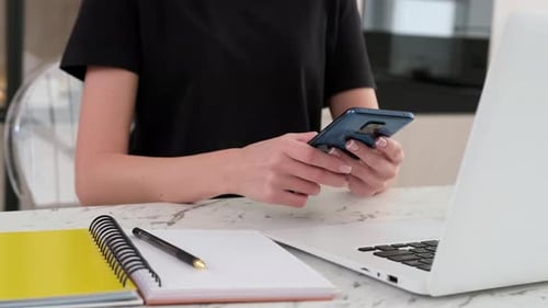Woman Using Phone at Desk with Laptop and Notepad