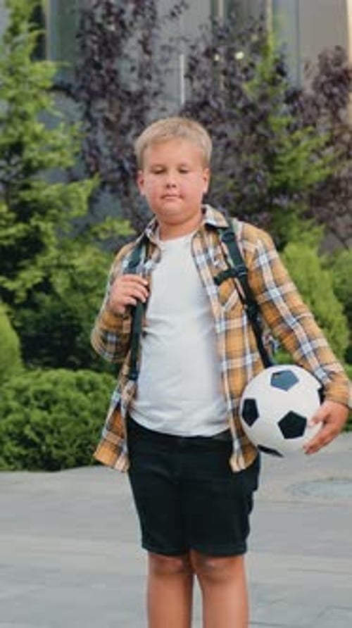 Portrait of Confident Blonde Boy Posing with Footbal Ball Outdoors Modern School Building Background