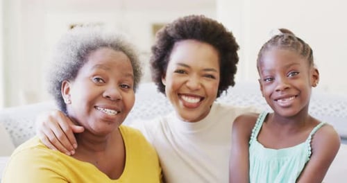 Portrait of smiling african american mother, daughter and grandmother embracing at home, slow motion