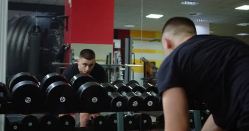 Medium Handheld Tracking In Gym Of Young Man Performing Bent Over Row With Dumbbell While Man Squats