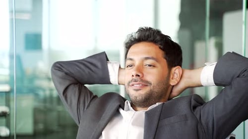 Close up. Young happy businessman in suit relaxes with hands behind head while sitting at workplace