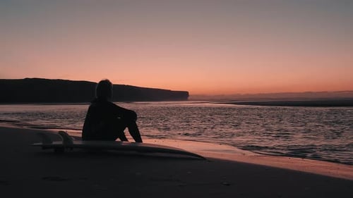 Close up shot of young woman surfer sitting on a sand near the ocean at golden sunset / sunrise.