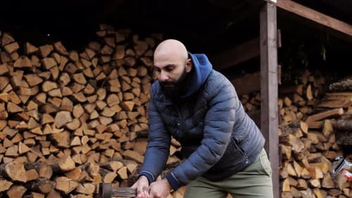 A Bearded Man is Chopping Firewood in the Backyard Near a Lot of Stacked Logs
