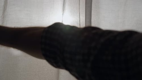 Hand reveals sun behind window curtains in early morning or sunset hour in city. Man pulls fabric