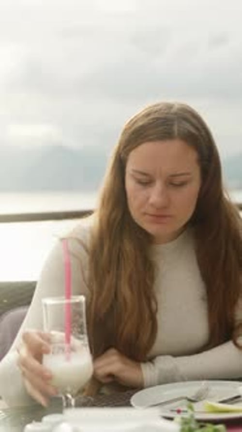 A young woman is drinking a cocktail in a restaurant by the sea, the sun is behind, and the glare on