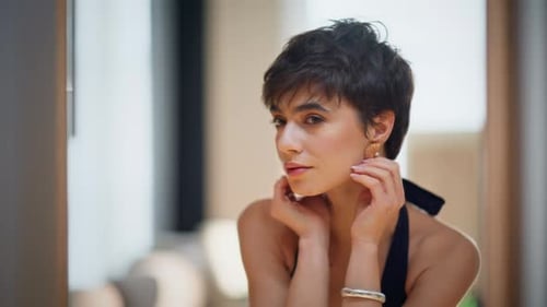 Attractive Woman Adjusting Gold Earrings in Mirror at Home