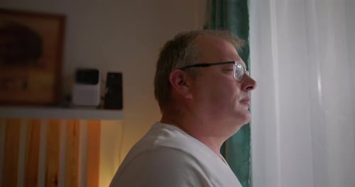Man gazing thoughtfully out of window, side profile view, glasses on, serene expression, reflecting