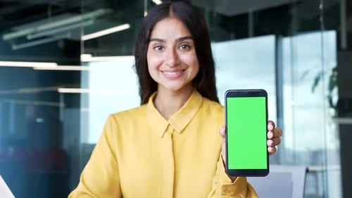 Happy businesswoman showing smartphone with green screen at workplace in office. Smiling female